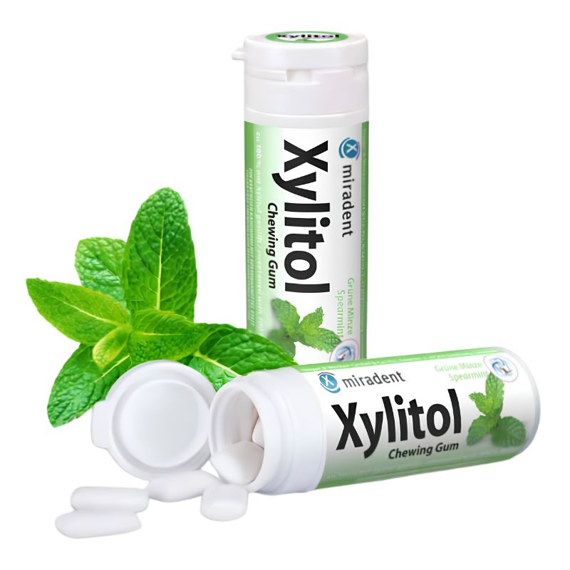 Two tubes of miradent Xylitol Chewing Gum, one standing upright and one lying on its side with the cap off, alongside a few white gum pieces and fresh green mint leaves on a white background.