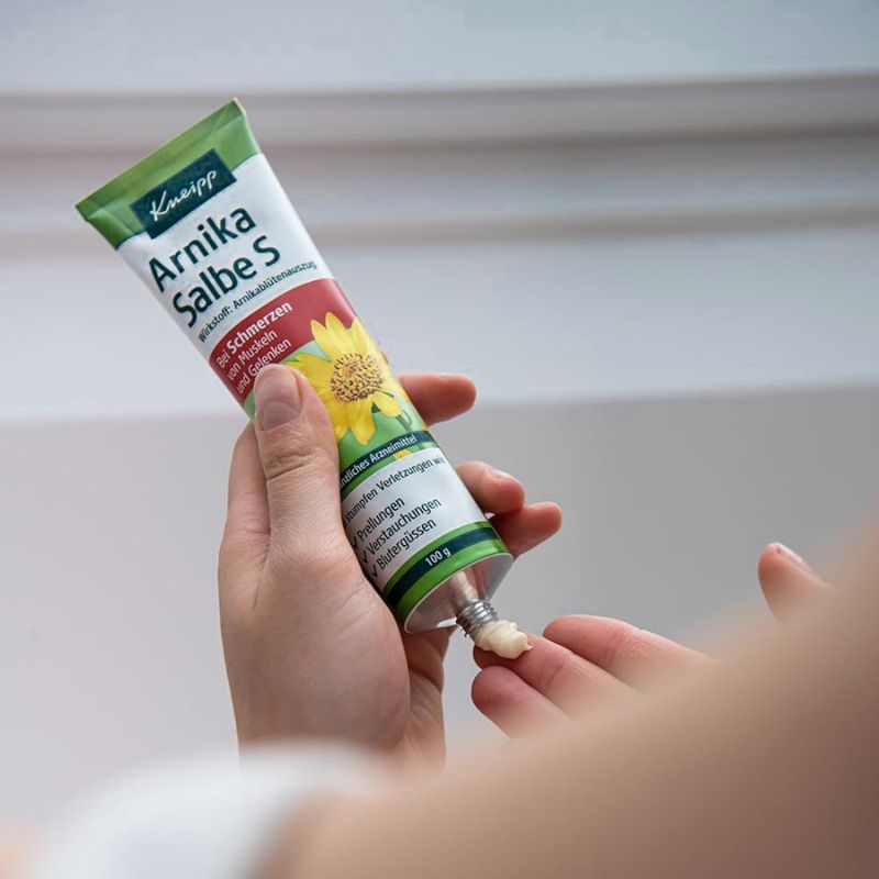 A hand holds a tube of Knipp Arnika Salbe cream, with another hand receiving a small dollop of the white ointment. The background is a soft, out-of-focus light gray interior wall.