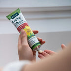 A hand holds a tube of Knipp Arnika Salbe cream, with another hand receiving a small dollop of the white ointment. The background is a soft, out-of-focus light gray interior wall.