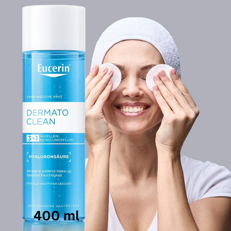 A bottle of Eucerin Dermato Clean facial cleansing fluid stands next to a smiling woman wearing a white head wrap and cotton pads on her eyes against a light gray background.