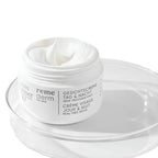 A white jar of Louis Richard Reme Derm dry cream with white cream visible inside, resting on a clear glass dish against a white background.