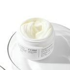 A white cosmetic jar of Yves Saint Laurent Reme Tagescreme moisturizer, filled with thick white cream and resting on a clear glass plate against a white background.