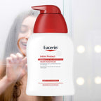 A bottle of Eucerin Intim Protect mild cleansing wash fluid with a red pump dispenser and white bottle, shown in front of a softly blurred background with a woman holding a mirror glass.