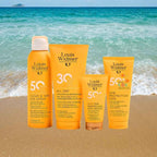 Four Louis Widmer sunscreen products in bright orange packaging are arranged on a sandy beach with turquoise ocean water in the background. The products include Clear & Dry Sun Spray, All Day Sun Protection, Face Sun Protection Anti-Aging, and Sun Protection Kids.