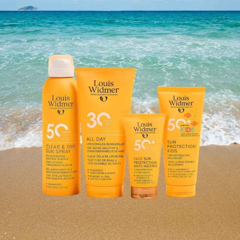 Four Louis Widmer sunscreen products in bright orange packaging are arranged on a sandy beach with turquoise ocean water in the background. The products include Clear & Dry Sun Spray, All Day Sun Protection, Face Sun Protection Anti-Aging, and Sun Protection Kids.