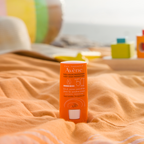 Avene sunscreen bottle on a beach towel with beach balls and sand toys in the background