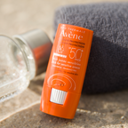 Orange Avene sunscreen stick on a textured surface with a glass bottle in the background